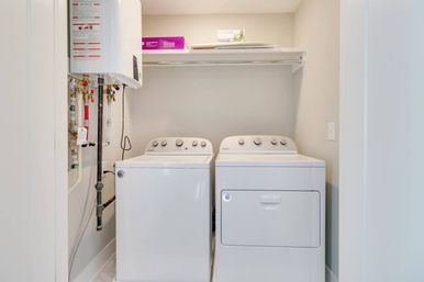 Bright compact home laundry nook with matching white washer and dryer side-by-side, overhead shelf, and tankless water heater with exposed plumbing.