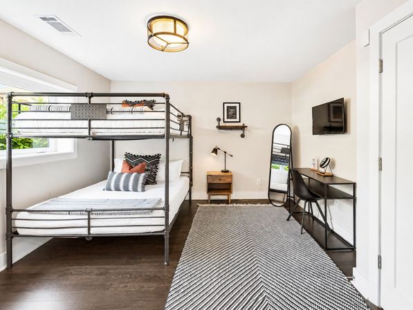 Bright, airy modern guest bedroom with industrial metal bunk bed (full bottom, twin top), wooden nightstand, black desk and chair, wall-mounted TV, full-length mirror, and chevron-pattern rug