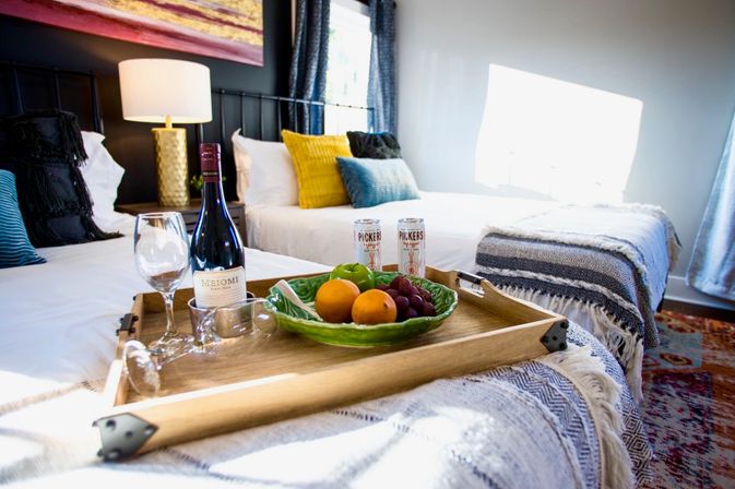 Sunlit modern guest bedroom with two beds, cozy throws and a wooden serving tray holding a bottle of red wine, two wine glasses, a bowl of fruit and two canned drinks.