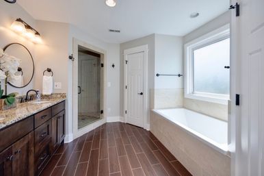 Bright spa-like master bathroom with dark wood double vanity and granite countertop, round mirror and sconces, glass-enclosed tiled shower, built-in soaking tub under a frosted window, and wood-look tile flooring.