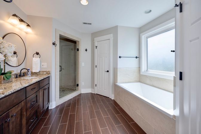 Bright spa-like master bathroom with dark wood double vanity and granite countertop, round mirror and sconces, glass-enclosed tiled shower, built-in soaking tub under a frosted window, and wood-look tile flooring.