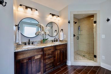 Sleek modern master bathroom with dark wood double vanity, granite countertop, two round mirrors, black fixtures, white orchid centerpiece, and tiled glass-enclosed walk-in shower.