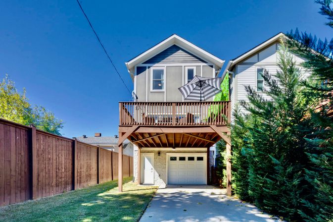 Two-story gray home with an elevated wooden deck and striped patio umbrella above a one-car garage, sunlit driveway, fenced yard and tall evergreens under a bright blue sky.
