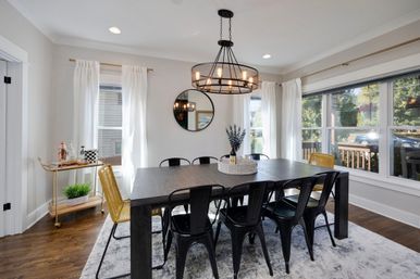 Bright modern farmhouse dining room with dark wood table, black metal chairs and yellow accent seats under a round chandelier, large windows with white curtains, hardwood floors and a gold bar cart.
