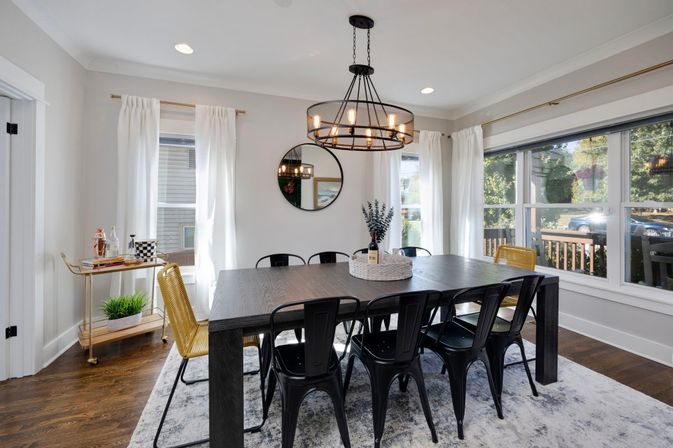 Bright modern farmhouse dining room with dark wood table, black metal chairs and yellow accent seats under a round chandelier, large windows with white curtains, hardwood floors and a gold bar cart.