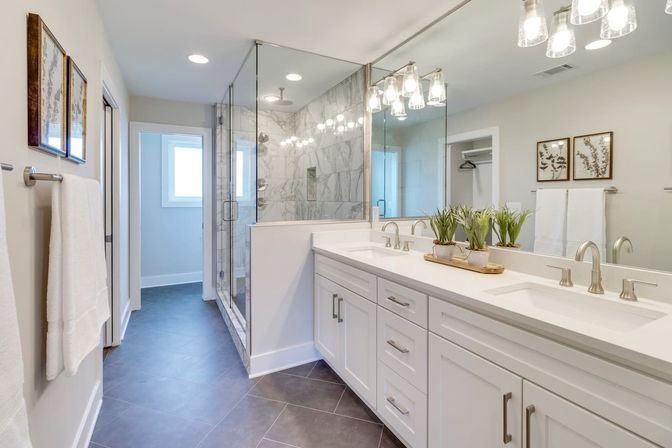 Bright, spa-like master bathroom with a white double-sink vanity and quartz countertop, dual brushed-nickel faucets, large mirror, glass-enclosed marble shower, gray floor tiles and a tray of small potted plants.
