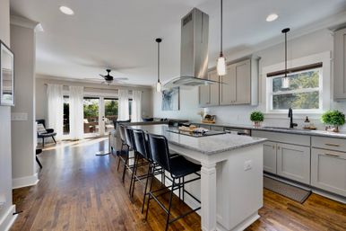Bright open-concept kitchen and living area with gray cabinets, granite island and black bar stools, stainless steel range hood, pendant lights, hardwood floors and sliding glass doors to a sunny backyard deck.