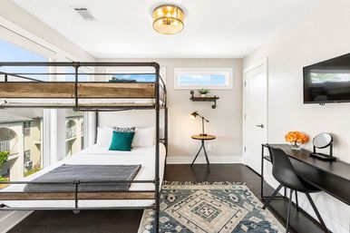 Bright, sunny bedroom with industrial metal-and-wood bunk bed, full lower bed with teal accent pillow, large windows showing blue sky, patterned rug, round bedside table with lamp, black desk and wall-mounted TV.