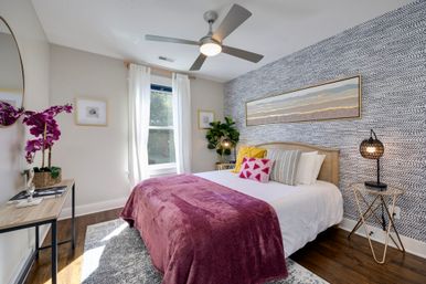Bright modern bedroom with patterned accent wall, queen bed in white linens topped with a plum velvet throw and colorful pillows, round ceiling fan, woven bedside lamp, potted plant, hardwood floors and sunlight streaming through a sheer-curtained window.