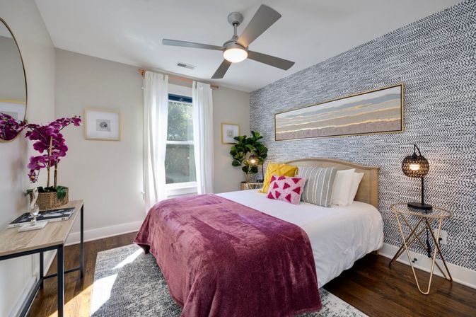 Bright modern bedroom with patterned accent wall, queen bed in white linens topped with a plum velvet throw and colorful pillows, round ceiling fan, woven bedside lamp, potted plant, hardwood floors and sunlight streaming through a sheer-curtained window.