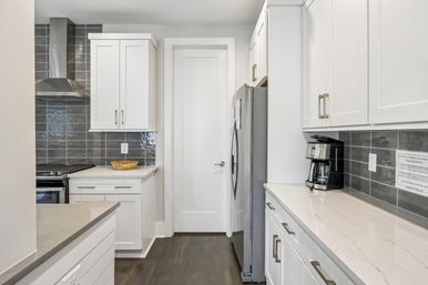 Modern white galley kitchen with shaker cabinets, gray subway tile backsplash, stainless steel range hood and refrigerator, white quartz countertops, coffee maker and wooden bowl on dark hardwood floors.