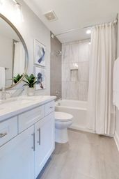 Bright, spa-like modern bathroom with white vanity and round mirror, potted plant on the counter, gray-tiled bathtub/shower with built-in niche, curved shower rod and white curtain.