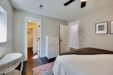 Sunny modern bedroom with neutral gray walls, dark hardwood floors, white wicker dresser and chair, colorful patterned rug, open walk-in closet and ceiling fan