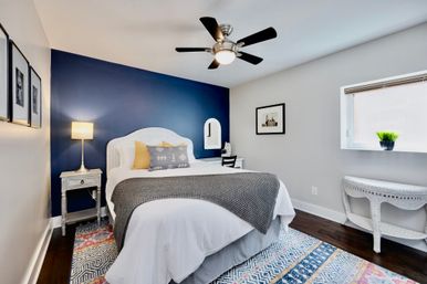 Cozy modern bedroom with a navy accent wall, white wicker headboard, queen bed dressed in white linens with a gray throw and yellow accent pillows, bedside lamp, ceiling fan, colorful rug and window plant.