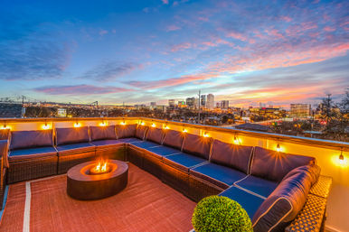 Rooftop patio at sunset with navy cushioned sectional, glowing string lights and a round fire pit overlooking a downtown city skyline.