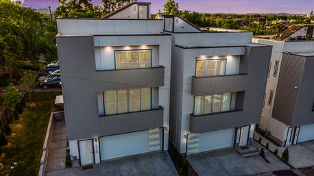 Aerial view of modern three-story townhomes with gray vertical siding, large windows and attached garages in a suburban neighborhood at dusk.