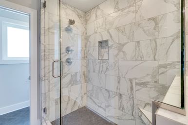 Bright modern walk-in shower with marble-look wall tiles, frameless glass door, chrome shower fixtures, recessed niche, and dark hexagon floor tiles.
