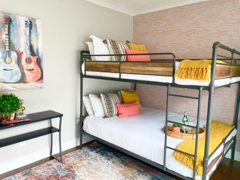 Cheerful modern guest bedroom with metal-frame bunk beds dressed in white linens, yellow throw blankets and colorful pillows; lower bunk features a wicker tray with a water bottle and glasses, guitar wall art above a black console table with a potted plant, textured pink accent wall and patterned area rug.
