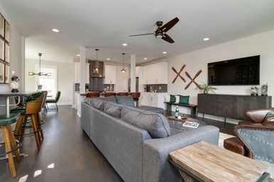 Open-concept living room and kitchen with roomy gray sectional, wood floors, white cabinets and dark tile backsplash, island with leather bar stools, ceiling fan, wall-mounted TV and crossed wooden oars wall decor.