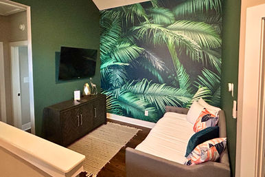 Cozy modern living nook with bold green palm-leaf wallpaper, dark wood media console and wall-mounted TV, gray daybed with colorful tropical pillows and woven rug