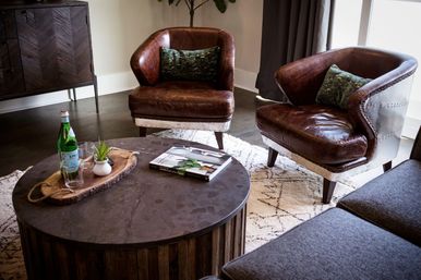 Cozy modern living room interior with two brown leather armchairs and green pillows, round rustic wood coffee table with bottle, glass, plant and book on a patterned rug — contemporary apartment lounge.