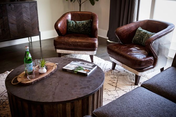 Cozy modern living room interior with two brown leather armchairs and green pillows, round rustic wood coffee table with bottle, glass, plant and book on a patterned rug — contemporary apartment lounge.