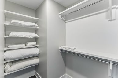 Bright modern home linen closet with white built-in shelves stacked with folded pillows and duvets, empty hanging rod and a neatly folded towel on a shelf.