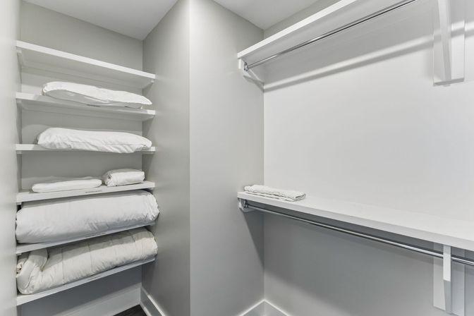 Bright modern home linen closet with white built-in shelves stacked with folded pillows and duvets, empty hanging rod and a neatly folded towel on a shelf.