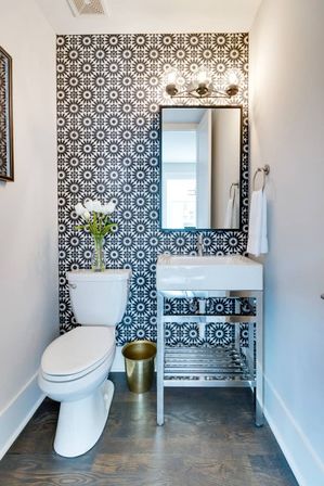 Chic small modern bathroom with black-and-white geometric floral wallpaper, white toilet topped with a vase of white tulips, chrome open-shelf vanity and rectangular sink with mirror, gold waste bin, and dark wood flooring.