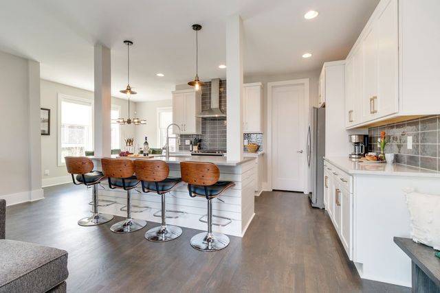 Sunlit modern open-concept kitchen with white cabinets, center island breakfast bar with four wood swivel stools, stainless steel appliances, pendant lights and dark hardwood floors.