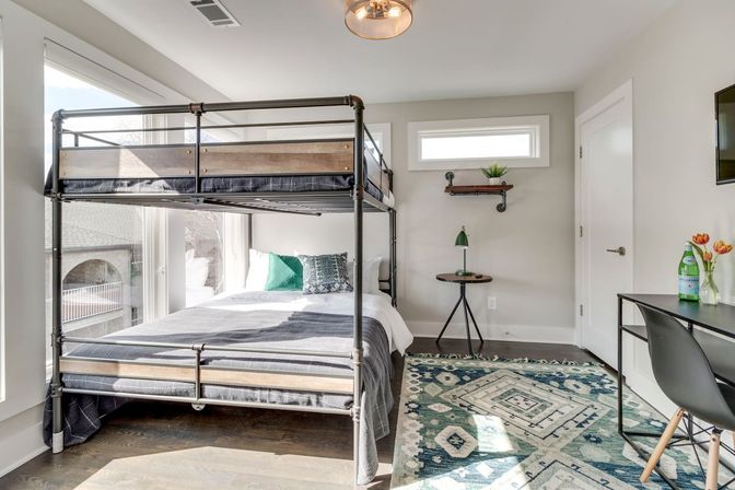 Sunlit modern bedroom with metal-frame bunk bed, teal geometric area rug, small round side table, compact desk with chair, and large windows letting in natural light.