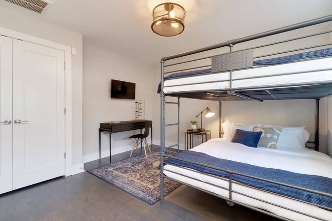 Cozy modern bedroom with metal bunk bed, lower bed dressed in white linens and navy throws, study desk with wall-mounted TV, bedside lamp and plant, gray floors and blue area rug.