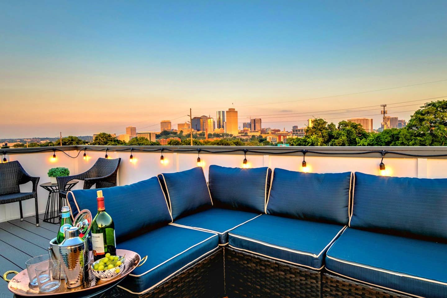 Cozy rooftop patio with deep-blue sectional sofa, string lights and a drinks tray, overlooking a glowing city skyline at sunset.