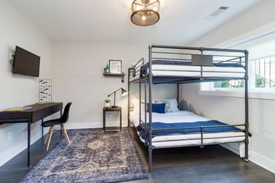 Bright modern bedroom with industrial metal bunk bed and navy blue bedding, patterned blue rug, black writing desk and chair, wall-mounted TV, bedside lamp and large window flooding the space with natural light.