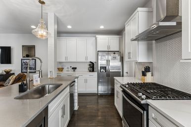 Modern open-concept kitchen with white shaker cabinets, quartz island with stainless undermount sink and spring faucet, stainless-steel fridge and gas range, glass pendant light, herringbone tile backsplash and dark hardwood floors.