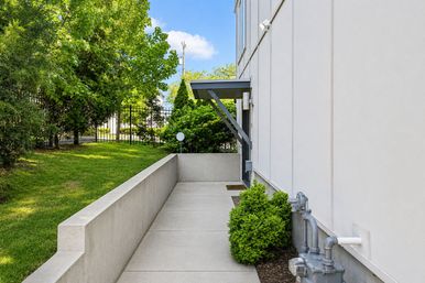 Sunny concrete walkway alongside a modern white building with a small metal awning over the side entrance, utility meters, trimmed green shrubs and a black iron fence with trees beyond