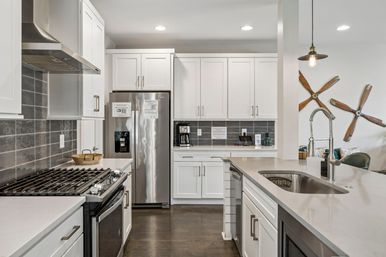 Bright modern kitchen with white shaker cabinets, stainless steel fridge and gas range, gray subway-tile backsplash, large island with stainless sink and spring-coil faucet, pendant light and playful wooden propeller wall art