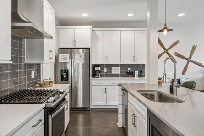 Bright modern kitchen with white shaker cabinets, stainless steel fridge and gas range, gray subway-tile backsplash, large island with stainless sink and spring-coil faucet, pendant light and playful wooden propeller wall art