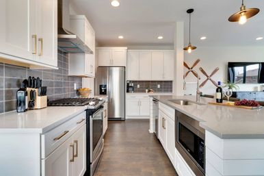 Bright modern open-concept kitchen with white shaker cabinets, stainless steel appliances, gray tile backsplash, and a large quartz island with pendant lights