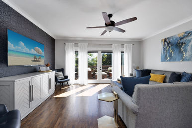 Sunlit modern living room with gray sectional and blue-yellow pillows, hardwood floors, ceiling fan, wall-mounted TV, and white curtains opening to a deck with outdoor seating