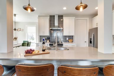 Bright modern open-concept kitchen with quartz island, mid-century wood bar stools, pendant lights, gray tile backsplash and a wine-and-cheese board on the counter.