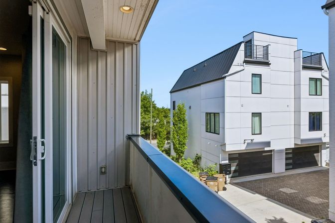 Second-floor balcony with sliding glass door overlooking modern white townhomes with rooftop decks, garages, and a clear blue sky