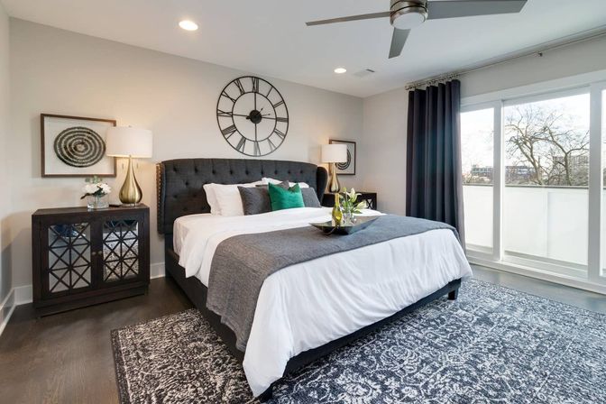 Bright contemporary master bedroom with tufted charcoal headboard, white bedding and gray throw, green accent pillows, oversized wall clock, mirrored nightstands with lamps, patterned rug, ceiling fan and sliding glass doors to a balcony with treetops.