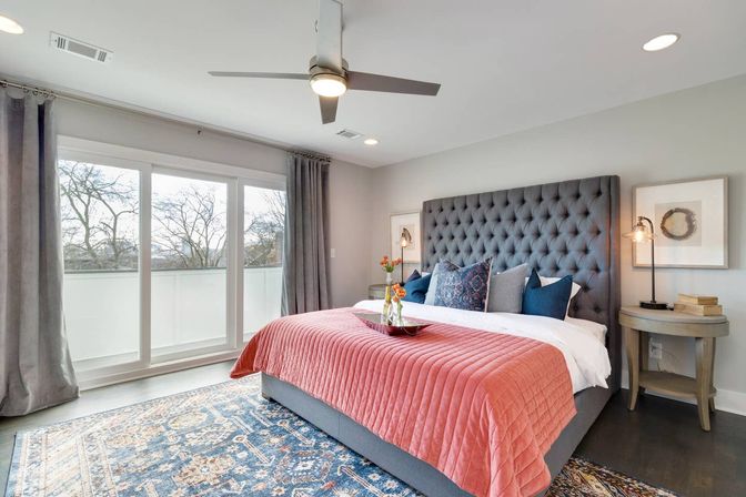 Light-filled modern master bedroom with tufted gray headboard, coral quilt on a king bed, patterned blue area rug, sliding glass doors with balcony view, gray curtains, ceiling fan and matching nightstands.
