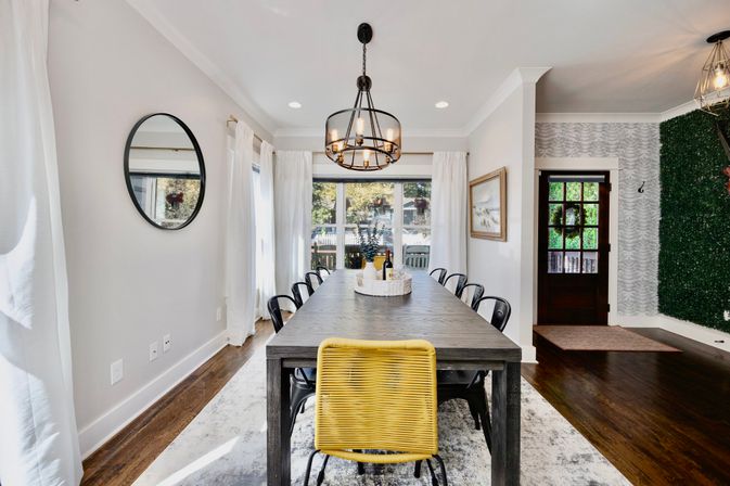 Bright contemporary dining room with a long dark wood table centered under an industrial chandelier, black metal chairs and a yellow woven accent chair, round wall mirror, white curtains, large windows, and hardwood floors.