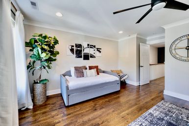 Sunlit modern guest bedroom with gray daybed and patterned pillows, hardwood floors, large potted fiddle-leaf fig, black ceiling fan, oversized wall clock and triptych wall art.