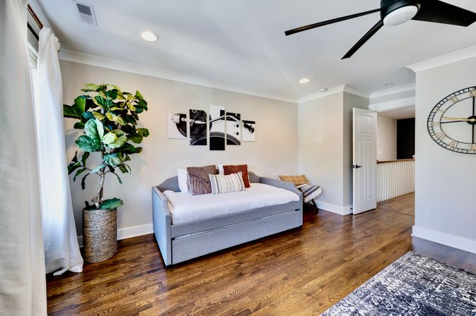 Sunlit modern guest bedroom with gray daybed and patterned pillows, hardwood floors, large potted fiddle-leaf fig, black ceiling fan, oversized wall clock and triptych wall art.