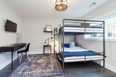 Bright modern guest bedroom with industrial metal bunk bed, white linens and navy throws, wall-mounted TV, compact black desk and chair, patterned blue area rug and a window flooding the room with natural light.