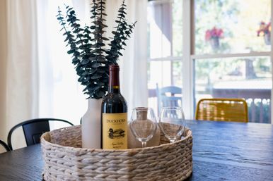 Sunlit dining table styled with a woven centerpiece tray holding a red wine bottle, two empty wine glasses and a white vase of eucalyptus, framed by large windows and patio chairs.
