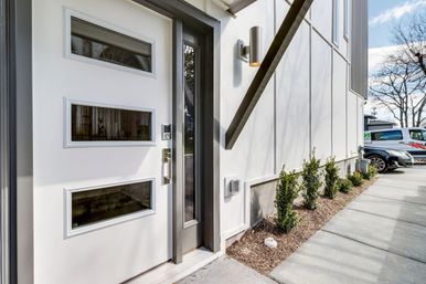 Modern white front door with three horizontal glass panels and smart lock, slim glass sidelight, wall sconce, neat shrub border along a concrete sidewalk and parked cars in the background.